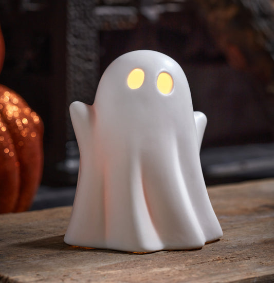 White ghost-shaped night light with glowing eyes on a wooden surface, blurred pumpkin in the background