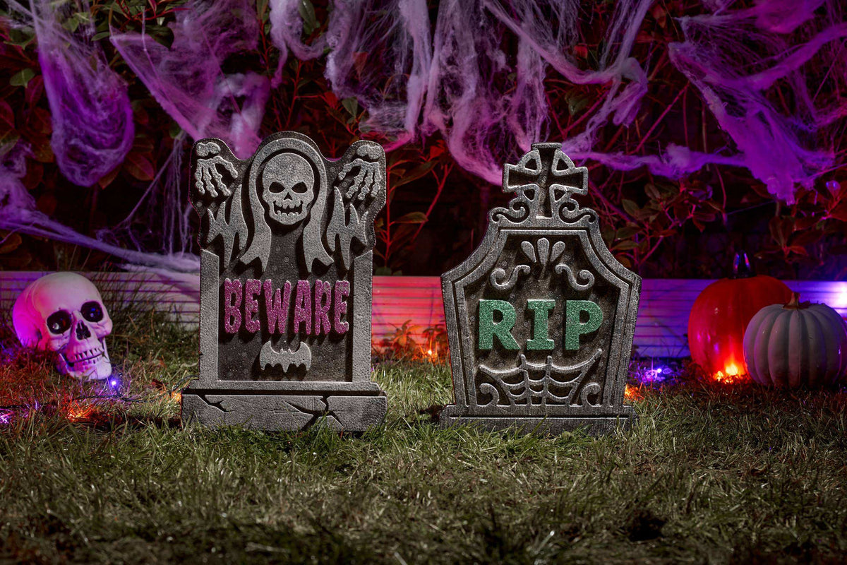 Two Halloween tombstone decorations on a grassy surface, one reading 'BEWARE' with a skull and bat motif, and the other reading 'RIP' with a cross and spiderweb design. The scene is styled with purple cobwebs, glowing pumpkins, and a skull prop in the background.