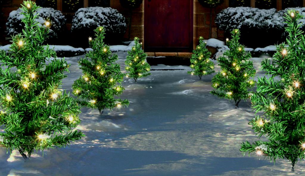 Decorative Christmas trees with lights on a snowy pathway leading to a door.