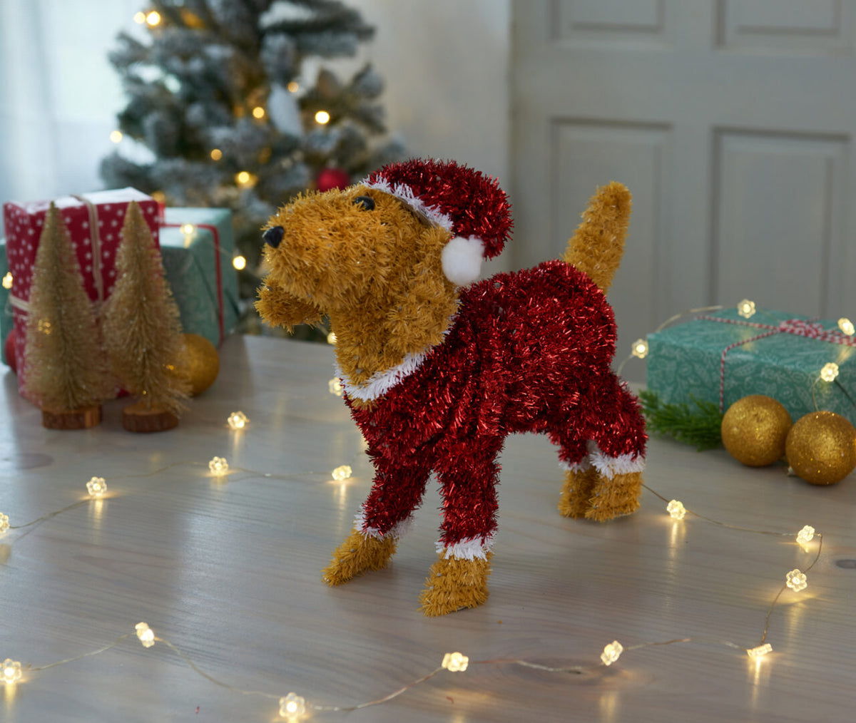 Decorative dog figurine in a red outfit with a Christmas tree and lights in the background.