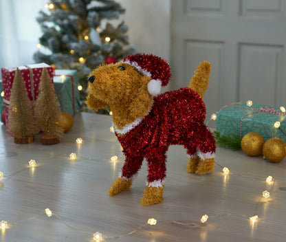 Decorative dog figurine in a red outfit with a Christmas tree and lights in the background.