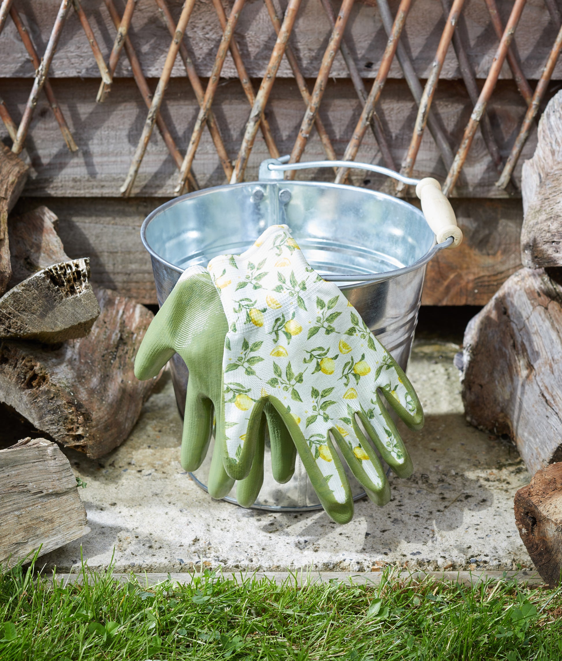 Green gardening gloves with floral pattern hanging on a metal bucket against a wooden fence background.