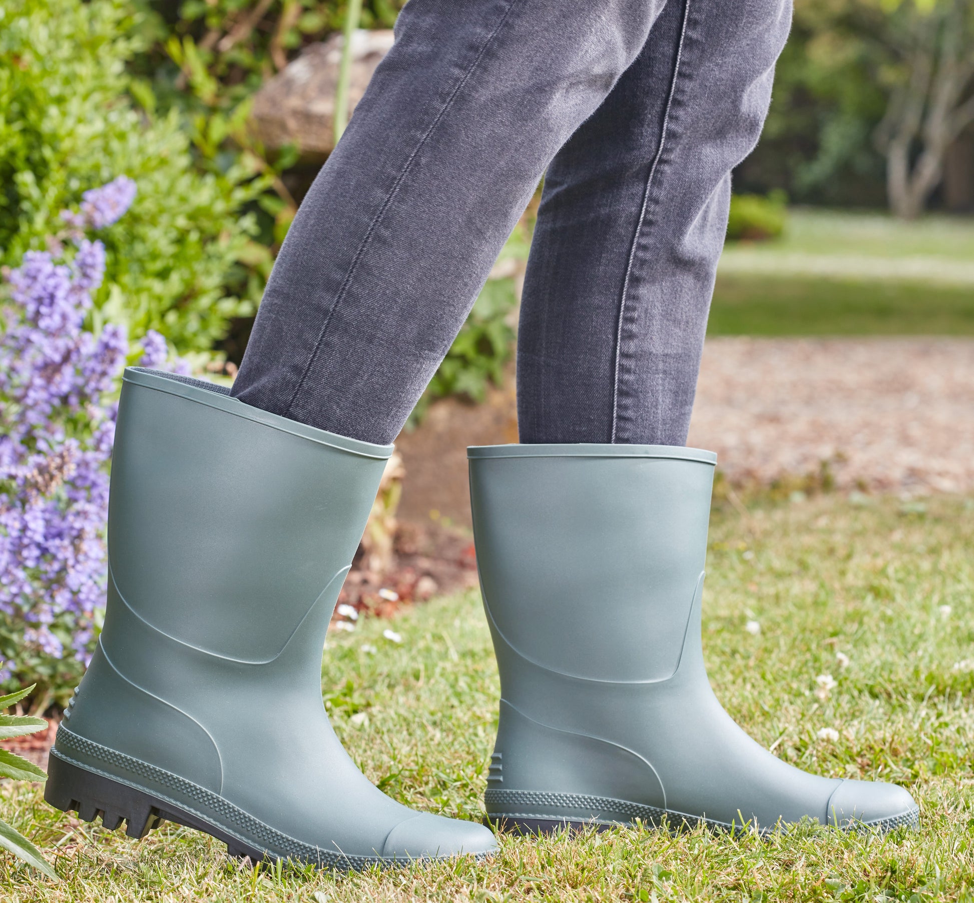 Person wearing green rubber boots on grass with a garden background