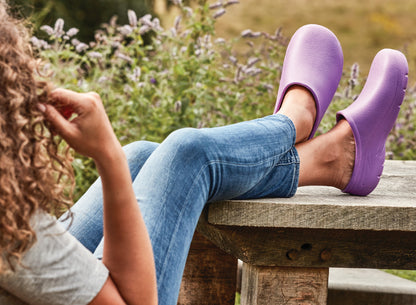 Person wearing purple shoes and blue jeans sitting on a wooden bench with a garden background