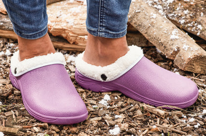 Purple clogs with white fur trim worn by a person on a wooden ground.