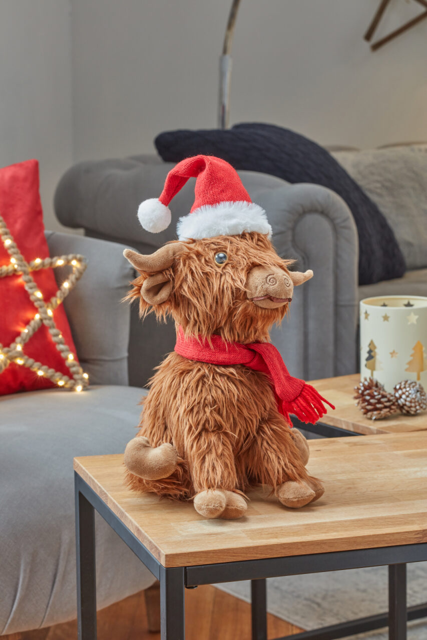 Plush toy Highland cow wearing a Santa hat and scarf on a wooden table.