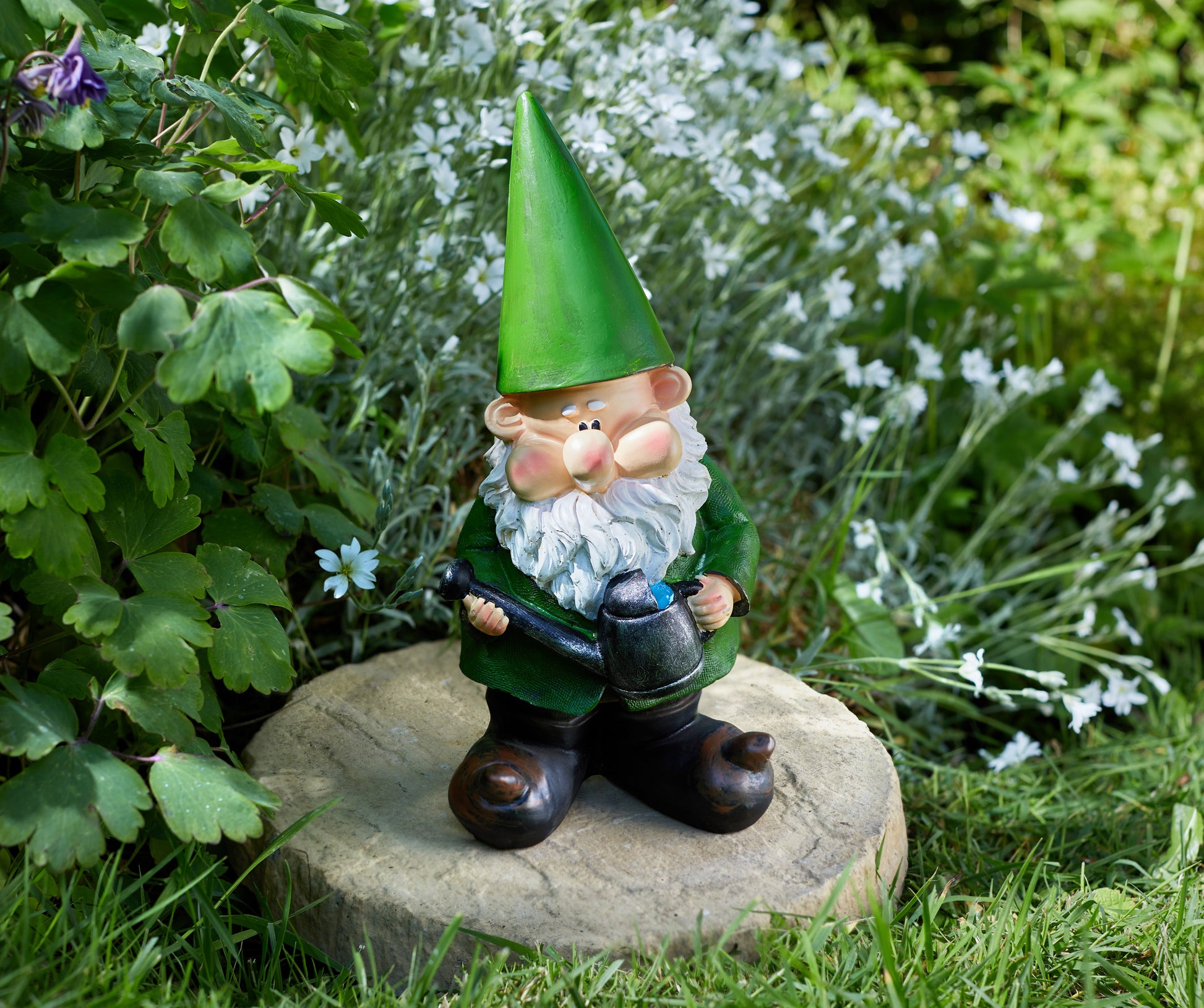Gnome figurine with a green hat and white beard holding a watering can, standing on a stone in a garden setting.