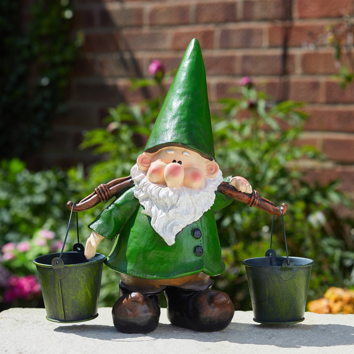 Gnome figurine with a green hat and holding two buckets against a brick wall and garden background.