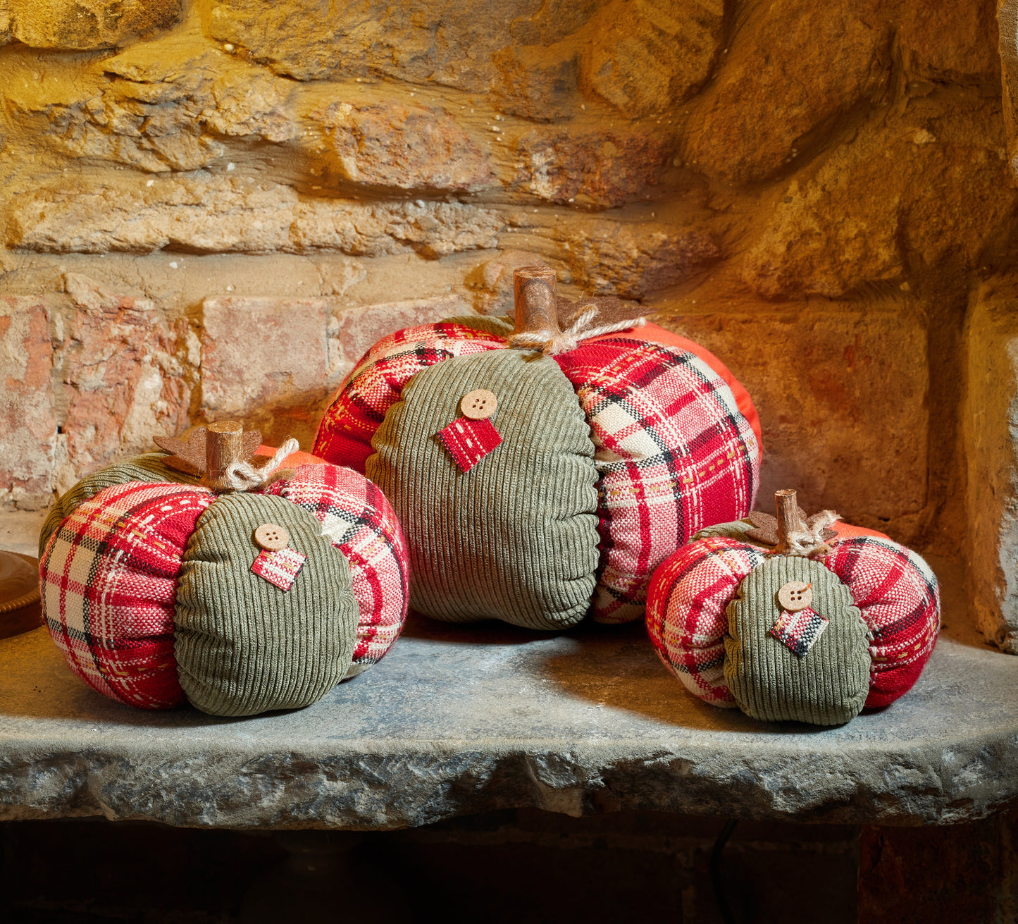Three fabric pumpkins in plaid and solid colors on a stone surface with a stone wall background.