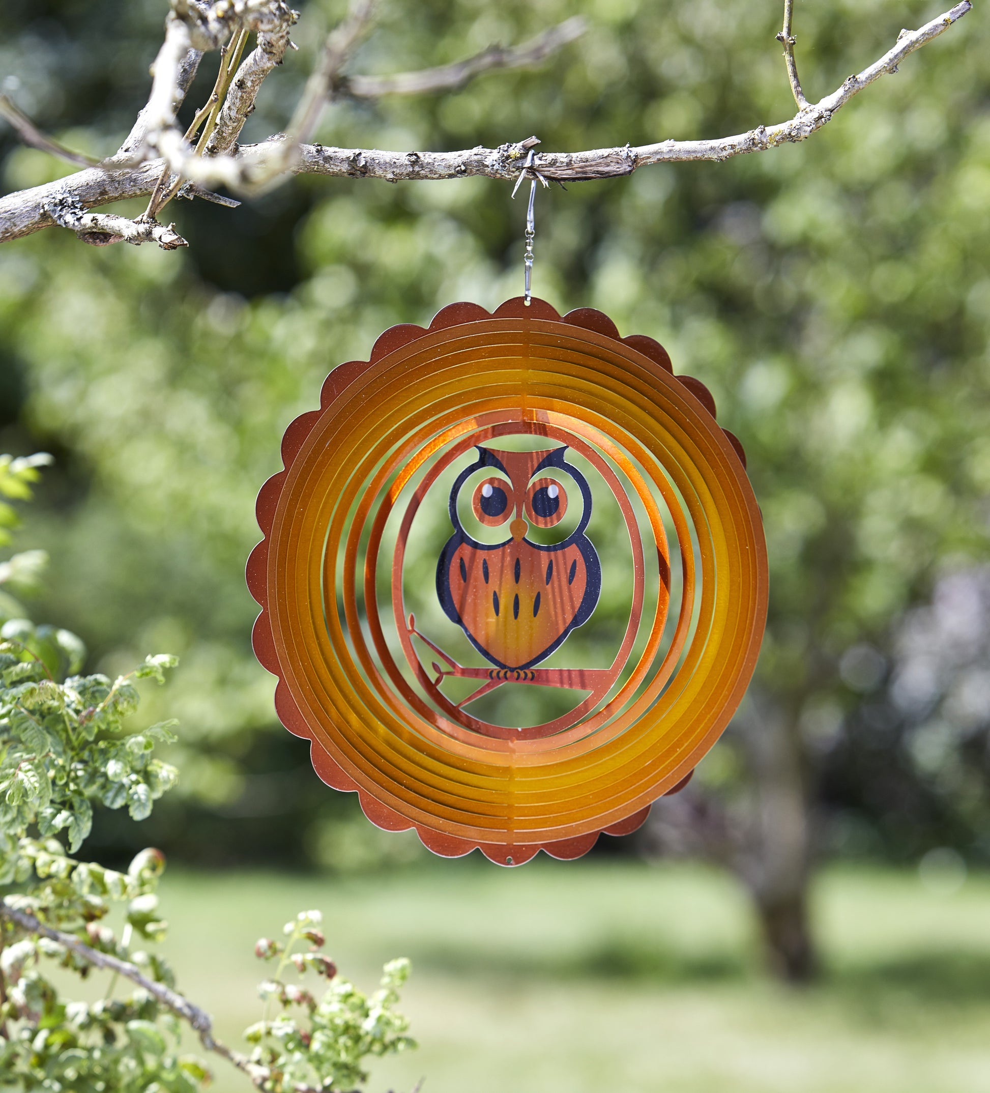 Decorative orange wind spinner with owl design hanging from a tree branch.