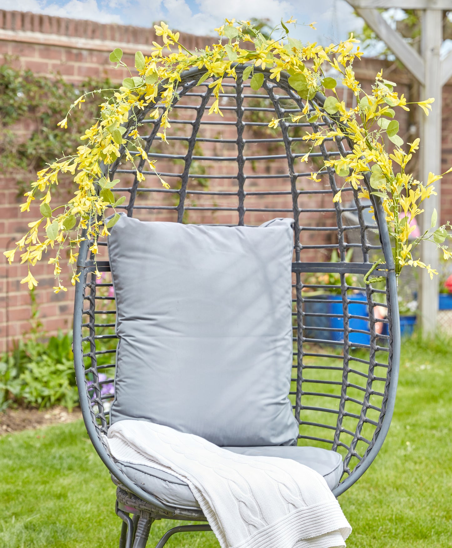 Egg-shaped wicker chair with a gray cushion and white blanket, surrounded by yellow flowers and greenery.