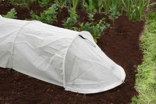 White garden tunnel cover on a bed of soil with plants in the background