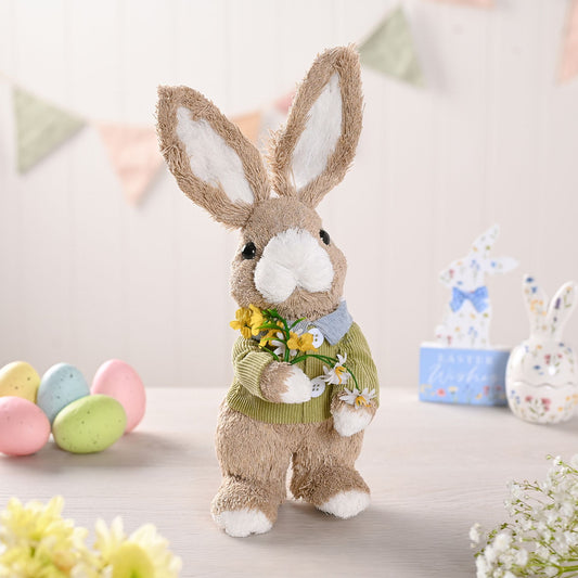 Plush bunny toy wearing a sweater with flowers, surrounded by Easter eggs and decorations.