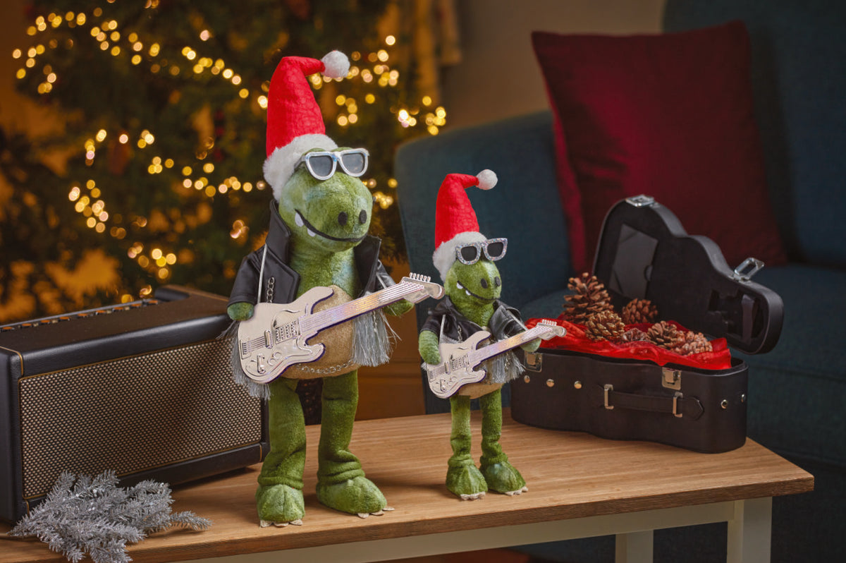 Two green dinosaur toys with Santa hats playing guitars on a table with a Christmas tree in the background.