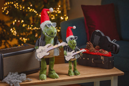 Two green dinosaur toys with Santa hats playing guitars on a table with a Christmas tree in the background.