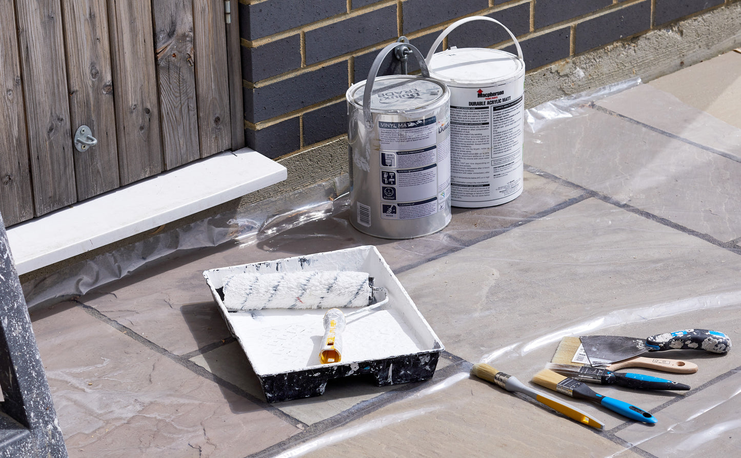 Paint cans, tray, and tools on a stone surface with a brick wall background