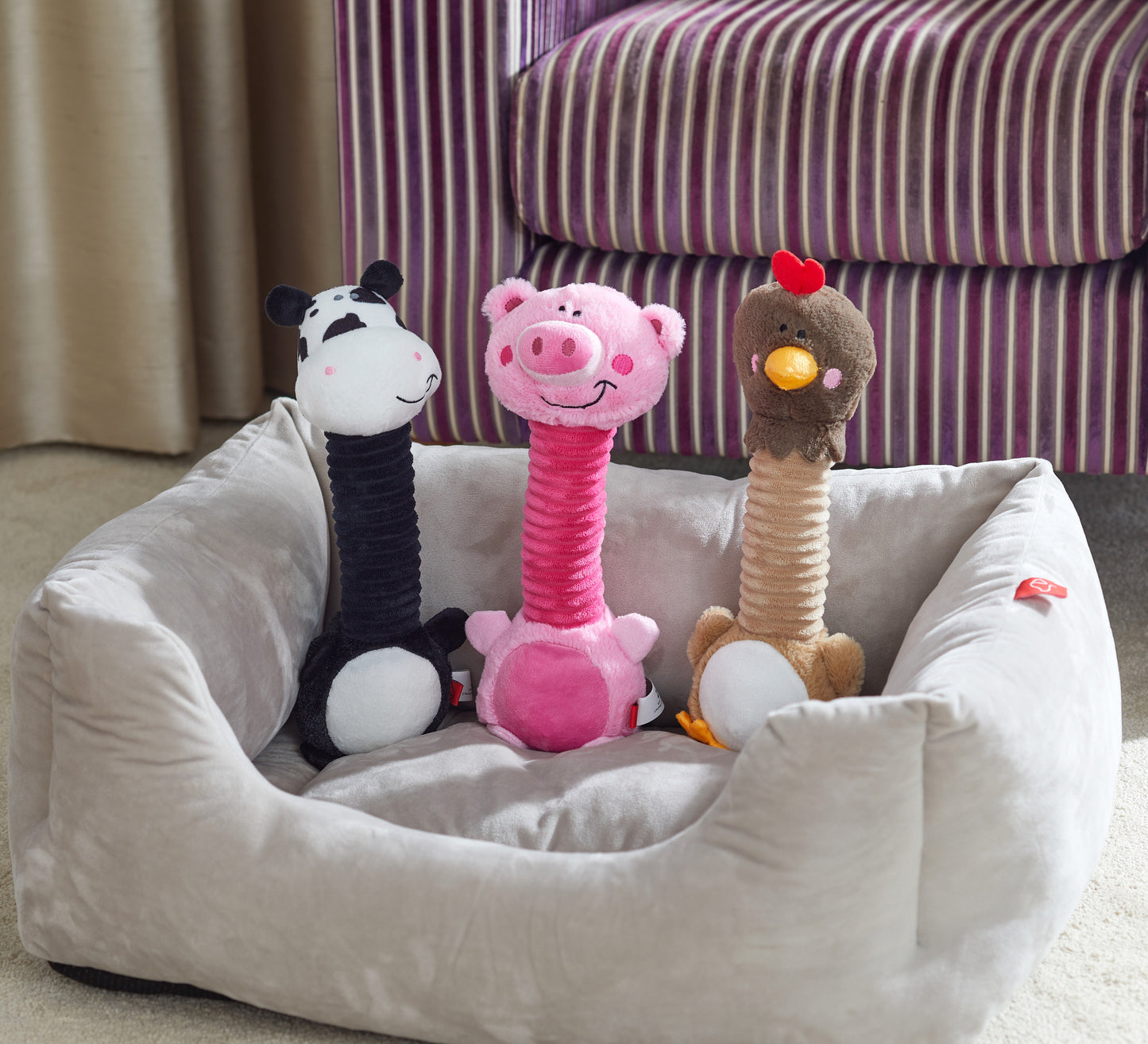 Three plush dog toys shaped like a zebra, pig, and monkey on a pet bed with a striped couch in the background.