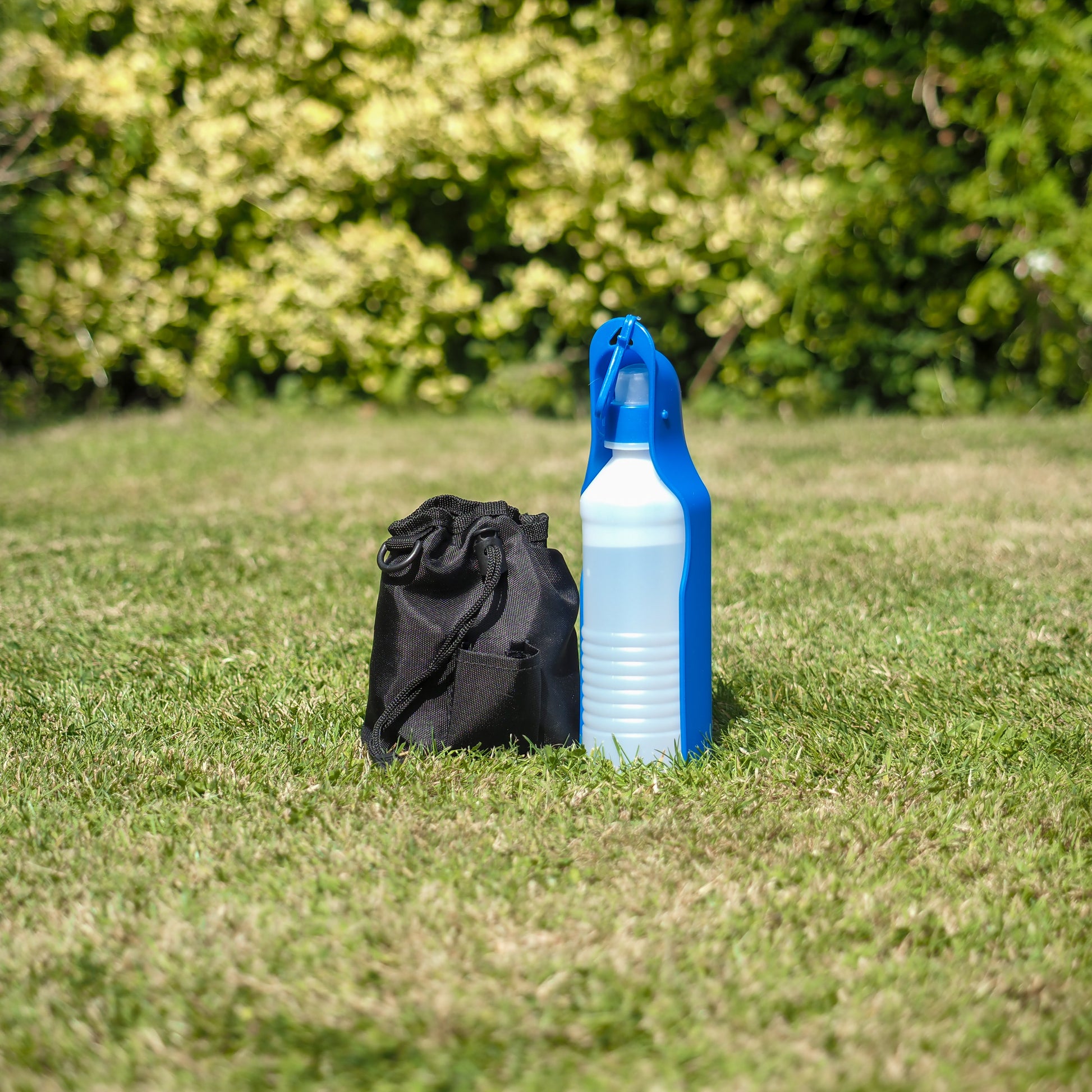 Black drawstring bag and blue and white spray bottle on grass with greenery in the background