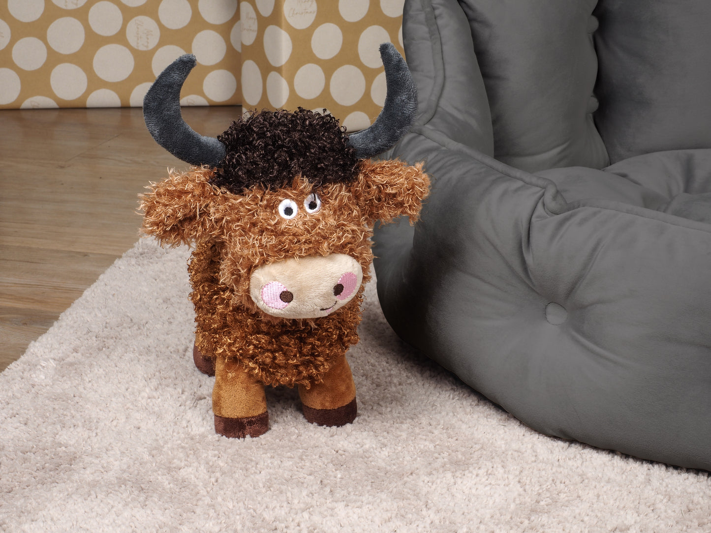 Plush toy cow on a carpeted floor next to a gray bean bag chair.