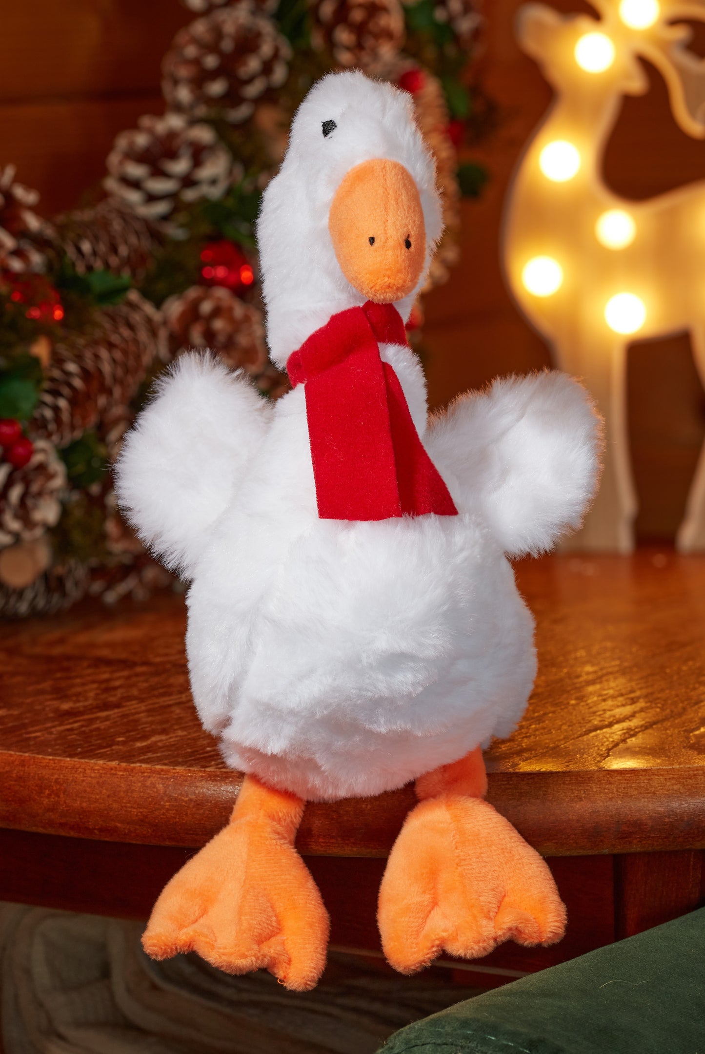 White plush duck with an orange beak and feet, wearing a red scarf, on a wooden surface with Christmas decorations in the background.