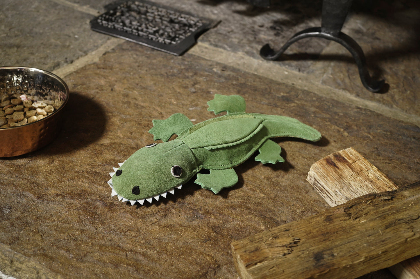 Green felt alligator toy on a stone surface with a bowl of treats and a piece of wood.