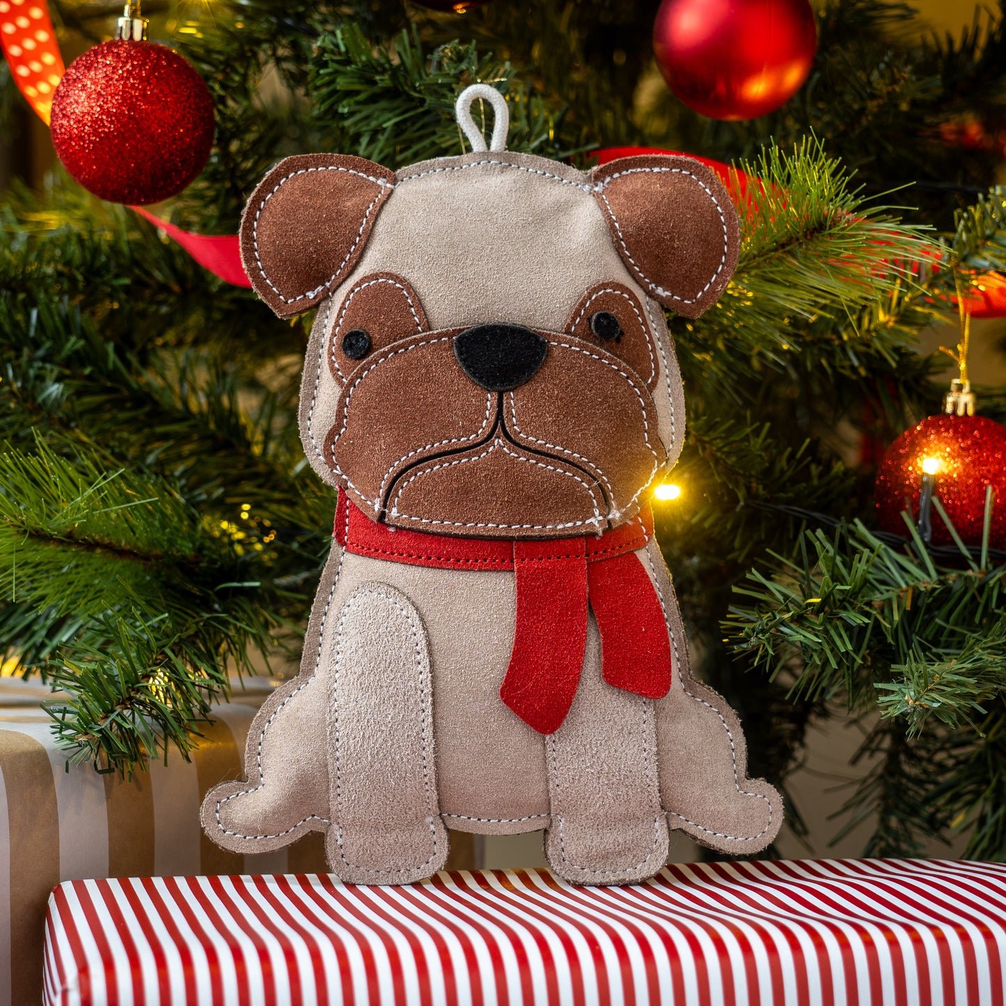 Dog-shaped Christmas ornament with a red scarf on a striped surface with a decorated tree in the background.