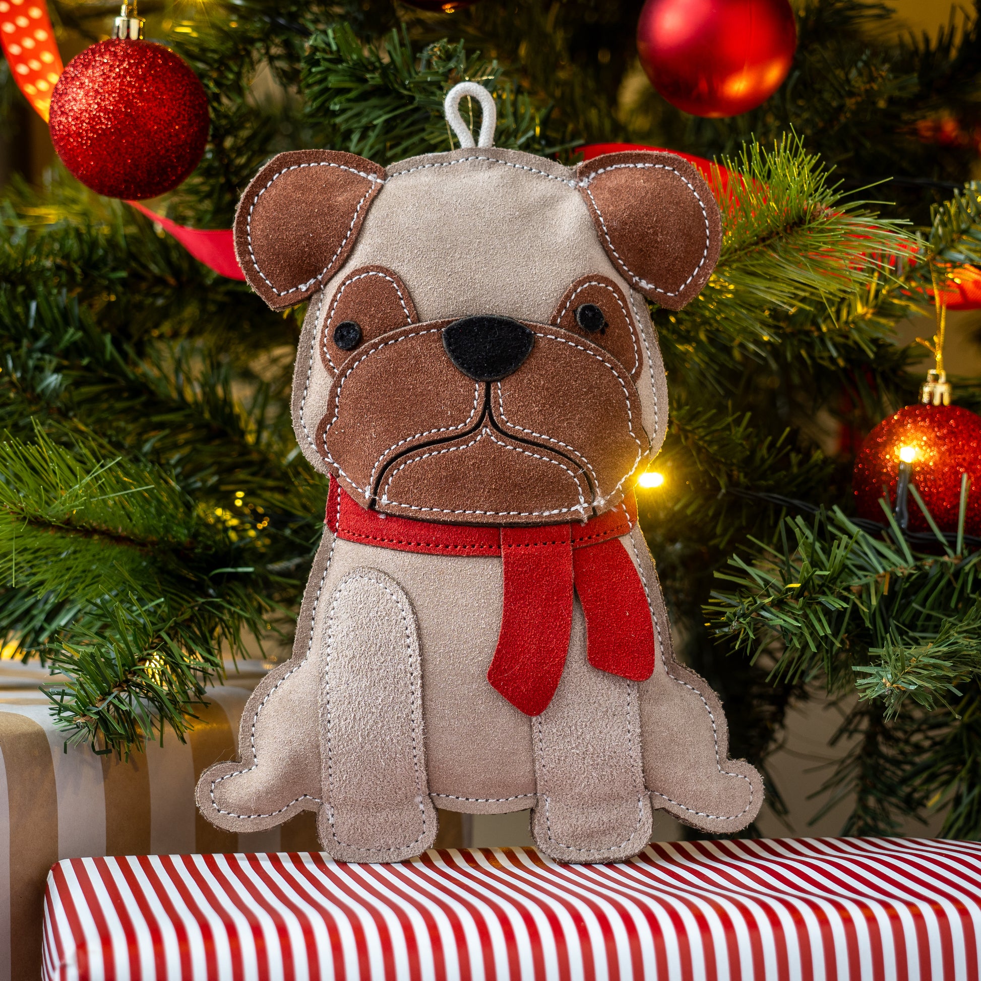 Dog-shaped Christmas ornament with a red scarf on a striped surface with a decorated tree in the background.