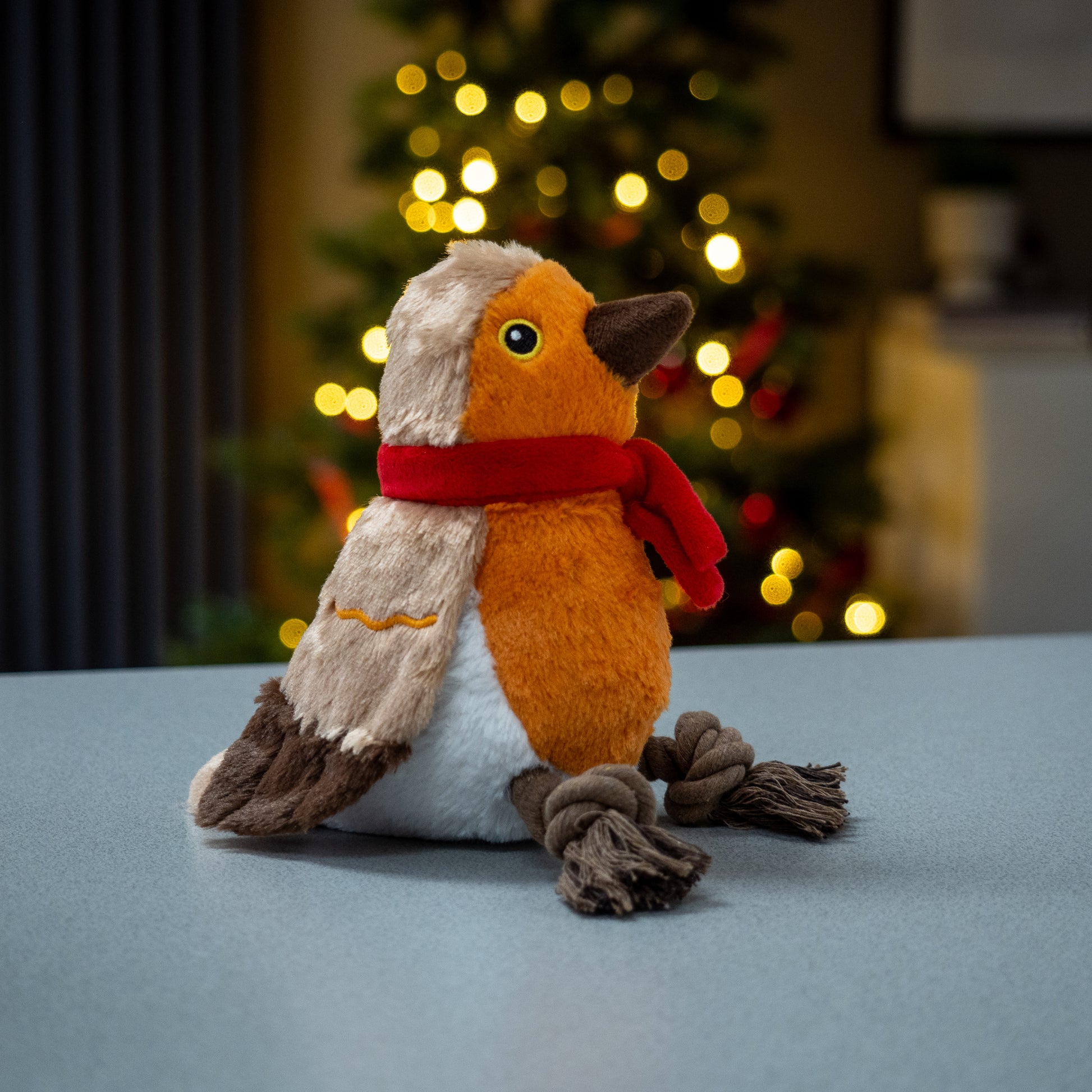 Plush toy resembling a bird with a red scarf in front of a blurred Christmas tree.