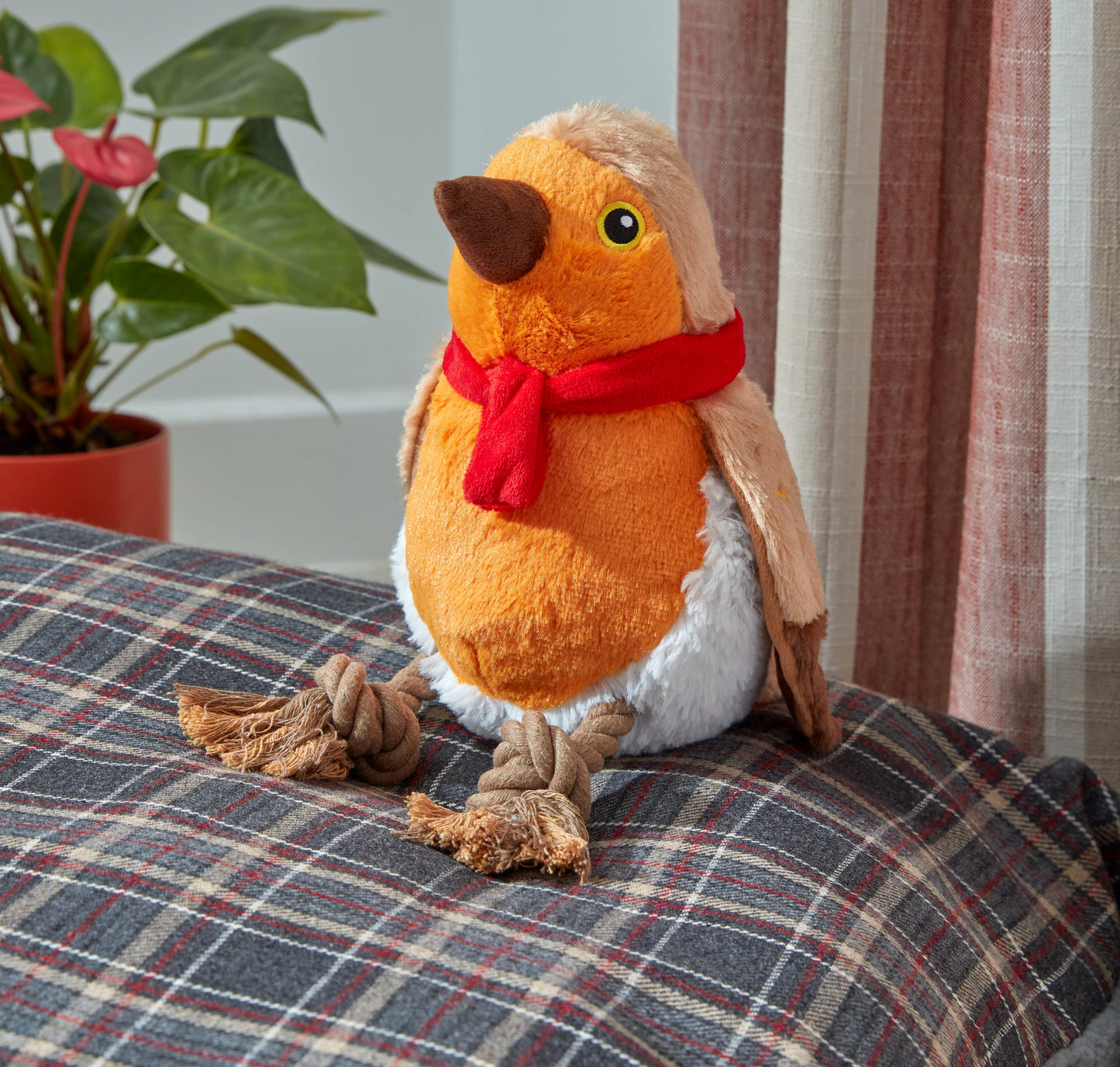 Plush bird toy with a red scarf on a checkered surface, with a plant and curtain in the background.
