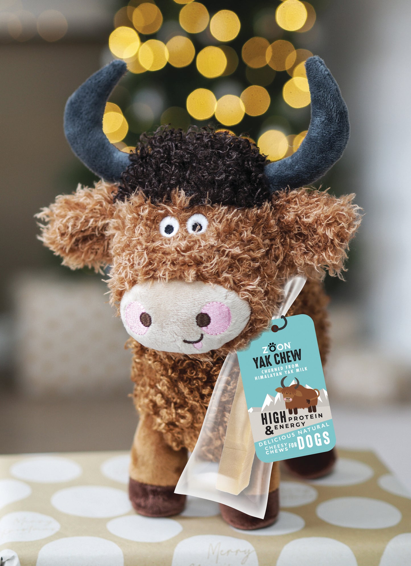 Plush toy yak with a tag on a patterned surface, blurred lights in the background