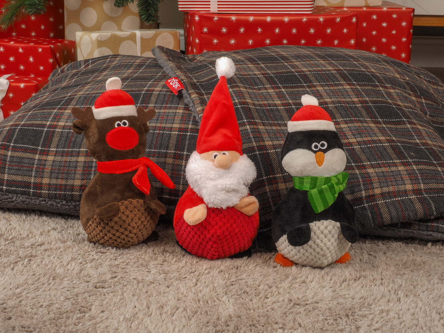 Three Christmas-themed plush toys (reindeer, gnome, penguin) on a plaid surface with presents in the background.