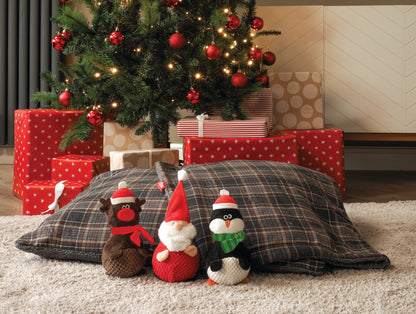 Three Christmas-themed plush toys on a plaid pillow in front of a decorated Christmas tree with presents.