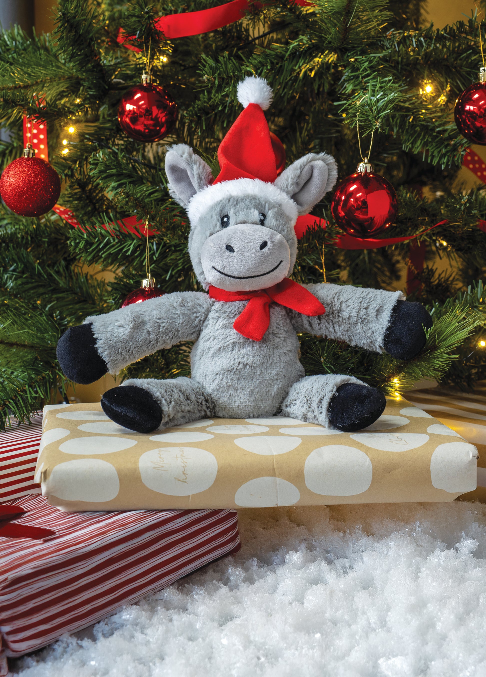 Plush donkey toy in a Santa hat and bow tie sitting on a wrapped present with a Christmas tree in the background.