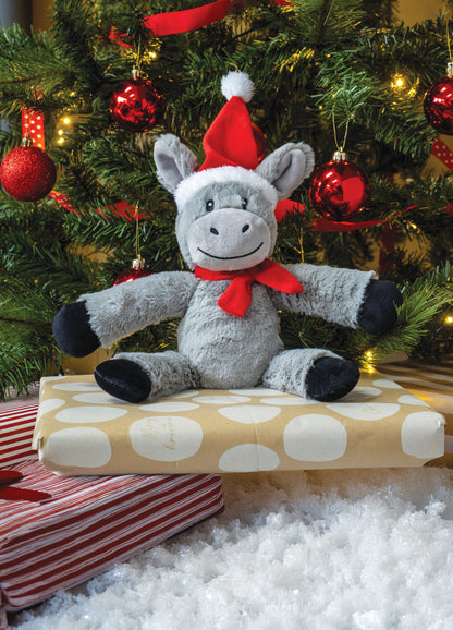 Plush donkey toy in a Santa hat and bow tie sitting on a wrapped present with a Christmas tree in the background.