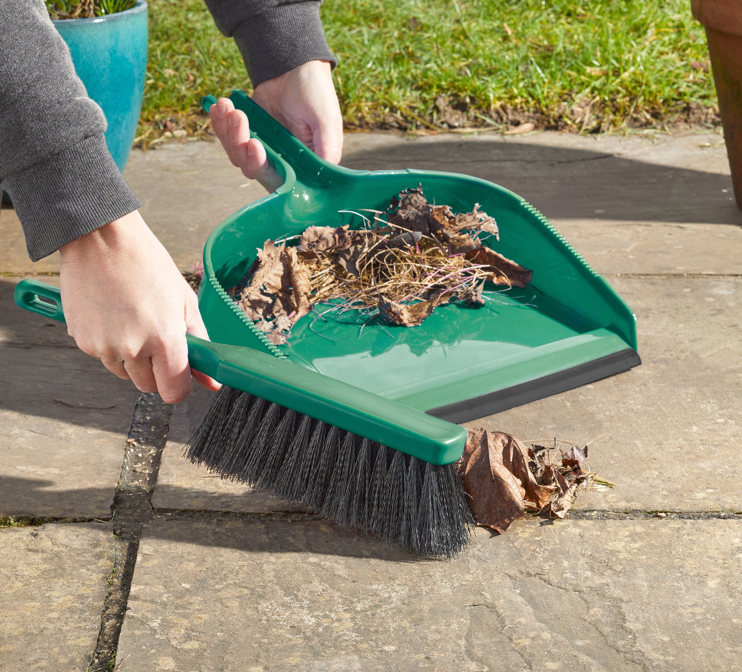 Person using a green dustpan and brush to collect leaves on a patio.
