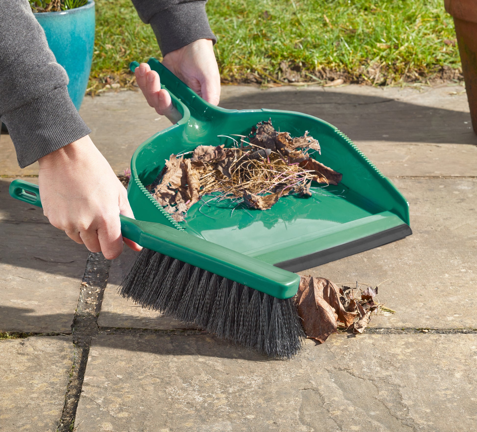 Person using a green dustpan and brush to collect leaves on a patio.