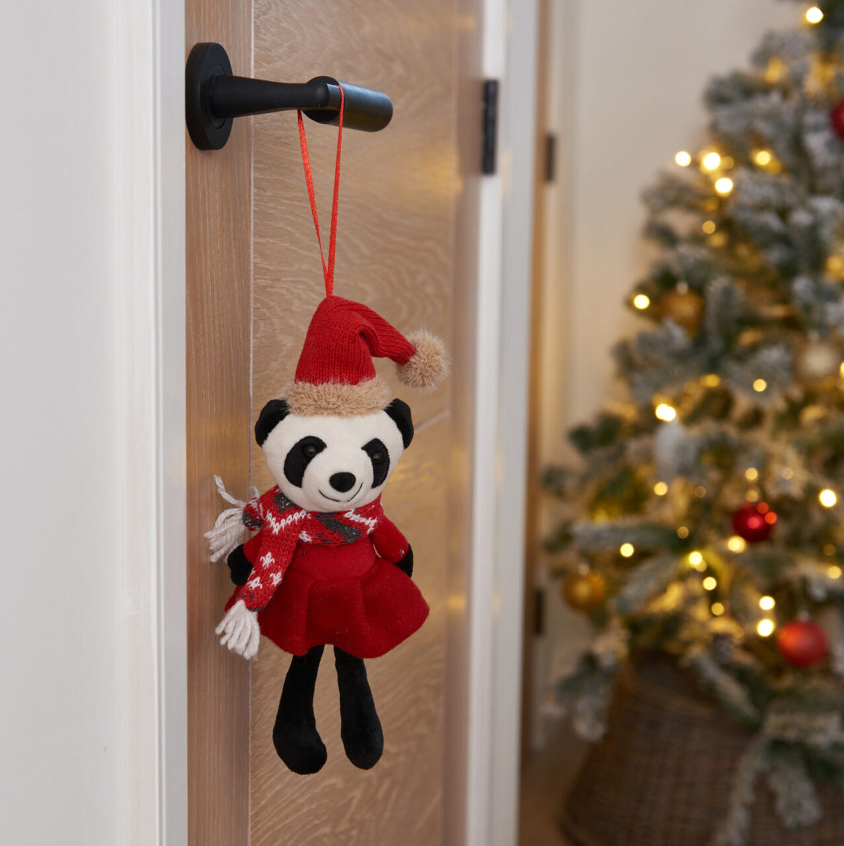 Panda toy in a red dress and Santa hat hanging on a door handle with a Christmas tree in the background.