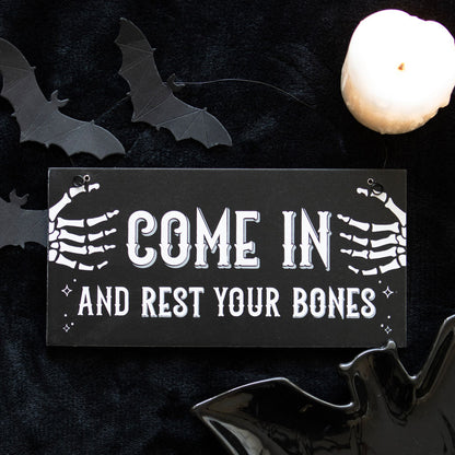 Halloween-themed sign with skeleton hands and text 'Come in and rest your bones' on a dark background with bats and a candle.