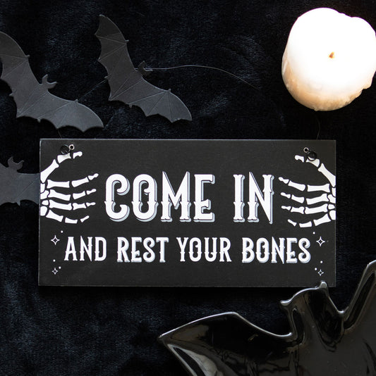 Halloween-themed sign with skeleton hands and text 'Come in and rest your bones' on a dark background with bats and a candle.