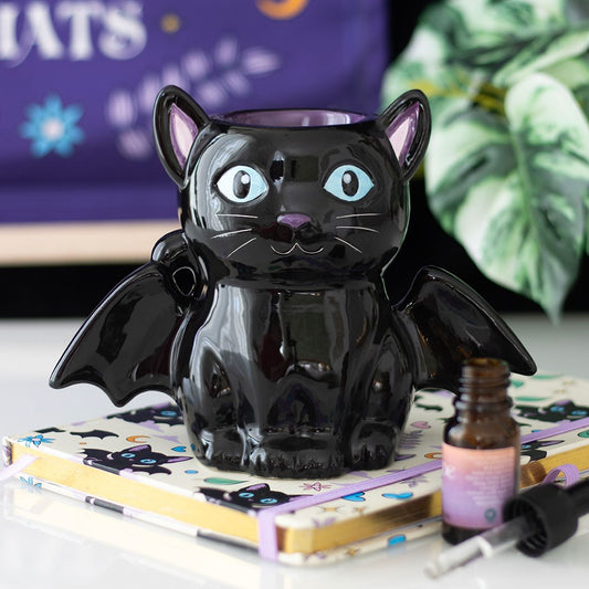 Black cat-shaped oil burner with bat wings on a surface with a notebook and bottle.