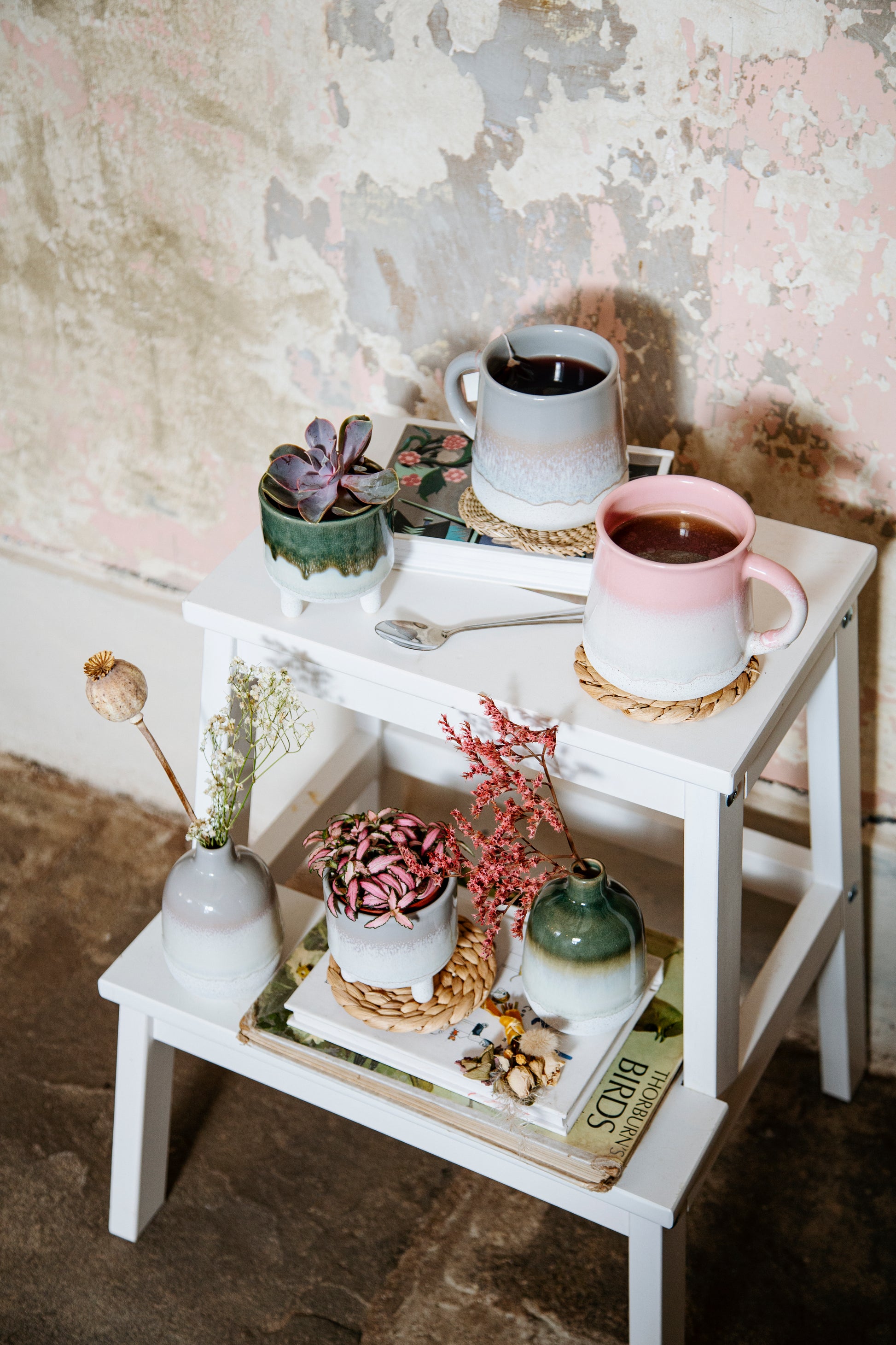 Small white table with decorative items including cups, plants, and books against a textured wall.