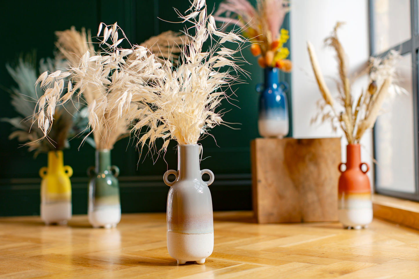 Small vase with dried plants on a wooden surface with other decorative vases in the background.