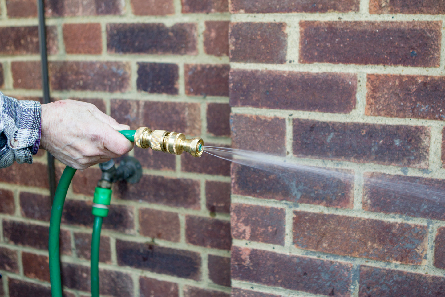 Person using a green garden hose with a brass nozzle against a brick wall.