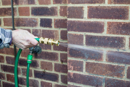 Person using a green garden hose with a brass nozzle against a brick wall.