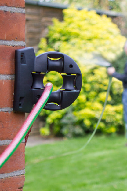 Wall-mounted device with a green ball attached, connected to a hose, against a brick wall and greenery background.
