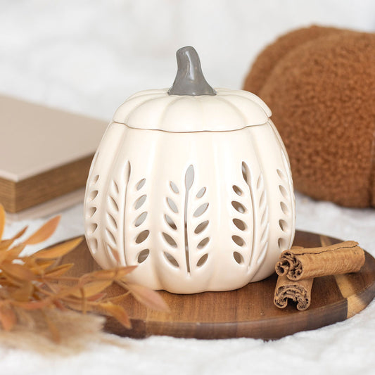 White pumpkin-shaped lantern with cut-out designs on a wooden surface with cinnamon sticks and leaves.