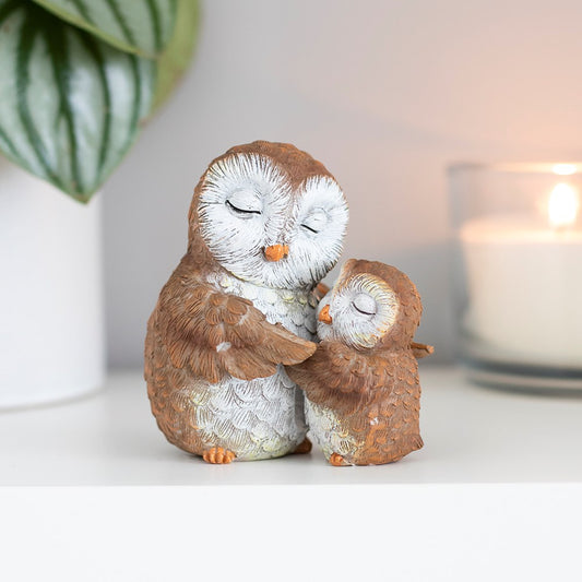 Two owl figurines embracing on a white surface with a blurred candle and plant in the background.