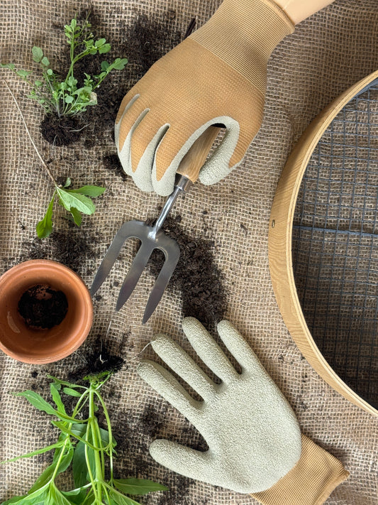 Gardening gloves, trowel, and small pot on a textured surface with soil and plants.