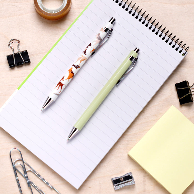 Two pens on a notebook with office supplies around