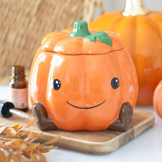 Pumpkin-shaped oil burner with a smiley face on a wooden board with pumpkins in the background.